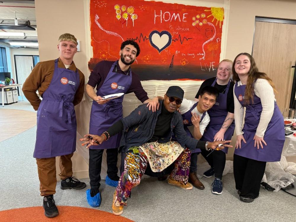 Volunteers in purple aprons laughing together in front of the 'Home' mural